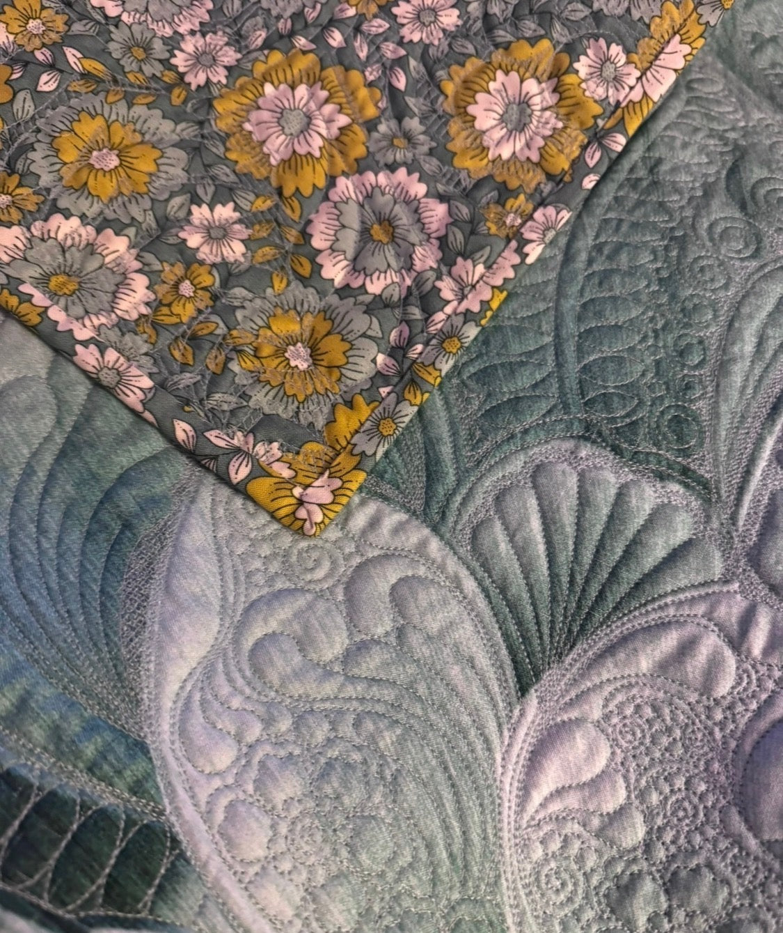 Close-up of dense feather longarm quilting in soft teal and green, with a grey and gold floral binding folded back at the corner